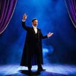 A professional male magician in a dark, elegant, modest tuxedo and a flowing cape, standing center stage. Dramatic spotlights in vibrant hues of deep blue and purple illuminate the scene, casting long shadows. Thick, atmospheric stage smoke billows around him, partially obscuring a large, ornate illusion prop. The magician is performing a subtle hand gesture, his expression focused and mysterious. The background is a grand, theatrical stage, with lush velvet curtains partially visible. Perfect anatomy, correct proportions, natural pose, well-formed hands, proper finger count. Professional photography, high detail, high quality, safe for work, appropriate content, fully clothed, family-friendly.